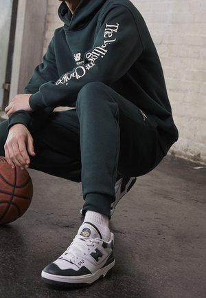 Persoon in een donkergroene hoodie en broek hurkt, houdt een basketbal vast, draagt witte en zwarte New Balance sneakers op een betonnen vloer.