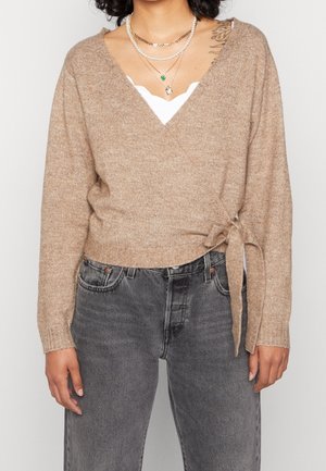 Femme portant un pull cache-cœur beige noué à la taille, un haut blanc à bords festonnés en dessous, un jean gris, et des colliers superposés en or et perles.