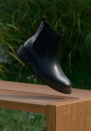 Bota de tornozelo em couro preto com painéis laterais elásticos flutuando acima de um banco de madeira, ao fundo folhagem verde.