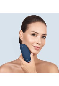 Blå ansiktsrengöringsapparat som hålls mot kinden, med en slät yta, böjd form och ett enkelt gränssnitt med minimala knappar.