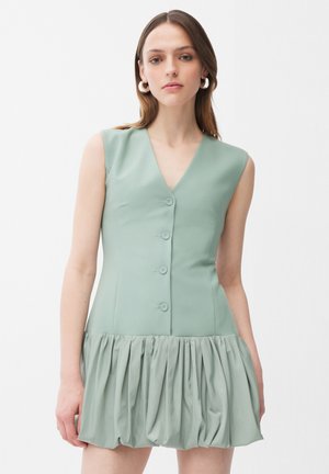 Mujer vestida con un vestido sin mangas de color verde salvia, con escote en V, botones en la parte delantera y falda fruncida, de pie frente a un fondo liso.