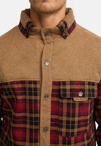 Bruine corduroy shirt met een rood en zwart ruitpatroon op de borst; voorzien van drukknoopsluiting en een flapzak aan de linkerkant. Textuurafwerking.