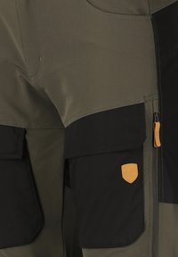 Vihreät ja mustat housut, joissa on suuret mustat cargo-taskut, joista yhdessä on pieni ruskea nahka-merkki ja vaaleanruskea vetoketjun nyöri.