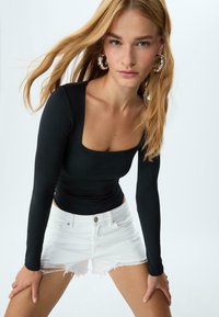 Mladá žena s dlouhými blond vlasy, která nosí stříbrné kruhové náušnice, černý dlouhý rukáv s čtvercovým výstřihem a bílé roztrhané šortky, pózující vpřed.