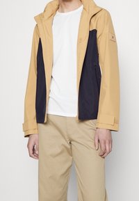 Tan og marineblå zip-up jakke med hætte. Har et to-farvet design, glat tekstur og justerbare ærmer. Bæres over en hvid skjorte.