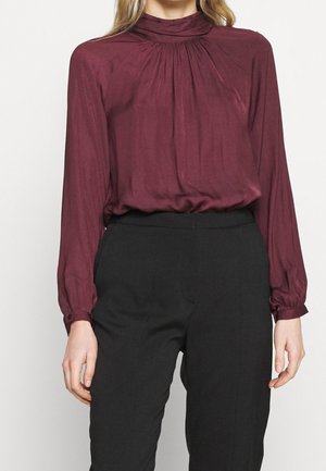 Bordowa bluzka z długim rękawem i wysokim kołnierzem, z plisami na ramionach i gładką, błyszczącą fakturą. W zestawie z czarnymi spodniami.