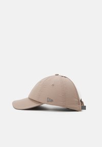 Casquette beige avec une visière courbée, fabriquée en tissu léger. Elle présente un logo discret sur le côté et de petits œillets de ventilation.