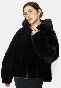 Veste zippée en fausse fourrure noire avec une grande capuche, des manches larges et une texture douce, dotée d'une fermeture éclair et d'une coupe décontractée.