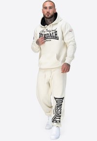 Crème hoodie och matchande joggers med svart tryckt logotyp. Har en frontficka, dragsnören och ribbade ärmslut. Vita sneakers fulländar looken.