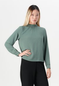 Ung kvinne med langt blondt hår iført en grønn bluse med høy hals og lange ermer, og svarte bukser, poserer med hånden på hoften mot en hvit bakgrunn.