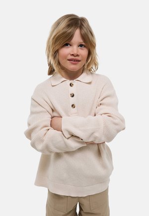 Enfant blond aux yeux bleus portant un pull en maille crème et un pantalon beige, debout les bras croisés sur un fond blanc.
