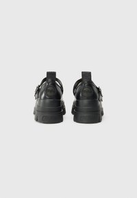Black platform sandals with a textured rubber sole, smooth finish, adjustable buckle, and circular logo detail on the heel.