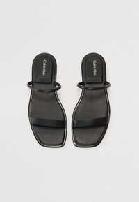 Juodos basutės su plokščiu padu, su dviem dirželiais: vienas horizontalus ir vienas apvalus akcentas viršutinei pėdos daliai. Calvin Klein logotipas ant pado.