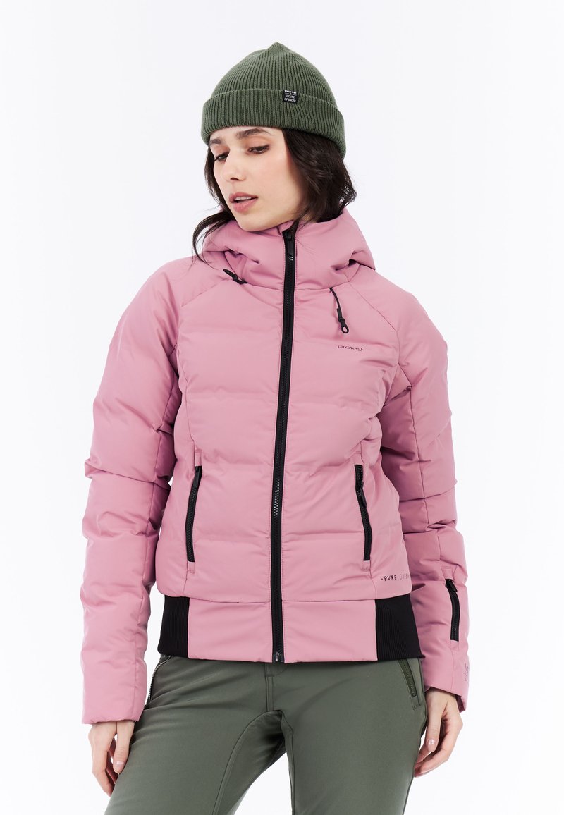Růžová naducaná bunda s černými zipy a manžetami, s kapucí a dvěma postranními kapsami. V kombinaci s olivově zelenou pletenou čepicí.
