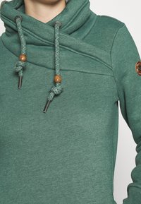 Felpa verde con un colletto alto e drappeggiato. Presenta lacci intrecciati con dettagli in legno e una toppa con logo circolare sulla manica.