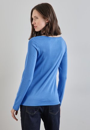 Langarmige blauwe top met een ronde halslijn en gladde textuur, gedragen met donkere spijkerbroeken, die een aansluitende silouet en minimalistisch ontwerp benadrukt.