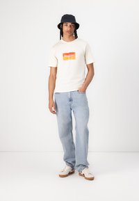 Crème kortemouwen t-shirt met een oranje logo, lichtblauwe denim jeans en witte sneakers met bruine accenten. Zwarte bucket hat.