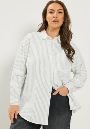 Vrouw met lang bruin haar draagt een los wit hemd met dunne zwarte strepen en een donkere broek, staand met één hand in de zak.