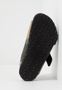 Infradito con suola nera texturizzata e tomaia con motivo mimetico, posizionati in verticale contro un angolo bianco su una superficie bianca.