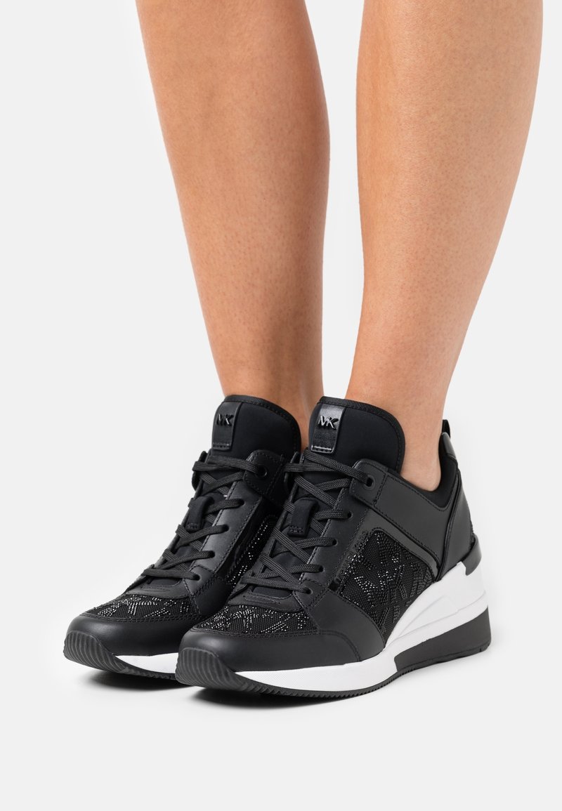 Baskets compensées noires et blanches avec détails en dentelle portées pieds nus sur fond blanc uni.