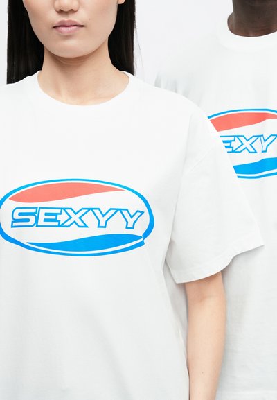 Bijele pamučne majice imaju upečatljiv grafički dizajn. Na prednjoj strani prikazuje se natpis "SEXYYY" u plavoj boji s crvenim i plavim naglascima u ovalnom obliku.
