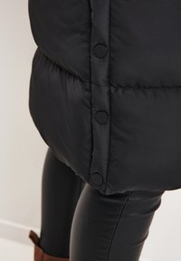 Veste matelassée noire avec un design matelassé, des boutons noirs et un tissu extérieur lisse. Portée avec des leggings noirs et des bottes marron.