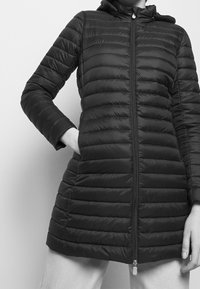 Zwarte gewatteerde jas met capuchon, voorzien van een ritssluiting, doorgestikte textuur en lange mouwen. Het ontwerp heeft zijzakken en een slanke pasvorm.