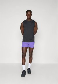 Homem musculoso em pé, vestindo uma t-shirt preta sem mangas, calções roxos, meias brancas e sapatilhas de corrida pretas, contra um fundo simples.