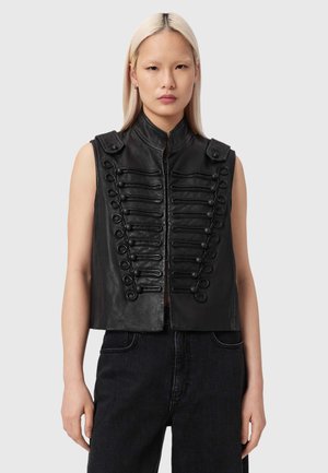 Vrouw met lichtblond haar, gekleed in een mouwloos zwart leren vest met decoratieve knooplusjes en een hoge kraag, gecombineerd met een zwarte broek.