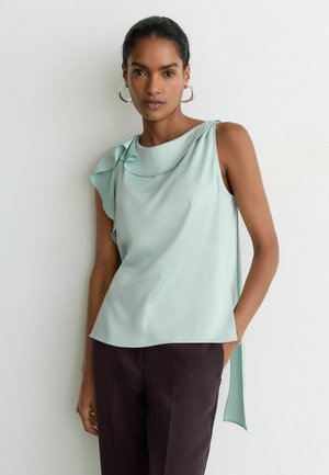 Femme aux cheveux foncés portant un haut bleu clair sans manches avec un détail à volants et un pantalon sombre, debout devant un mur blanc uni.