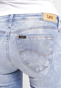 Vaqueros de mezclilla azul claro con un acabado desgastado, que presentan un parche de cuero con el logo de Lee y un detalle distintivo de costura de bolsillo curvado.