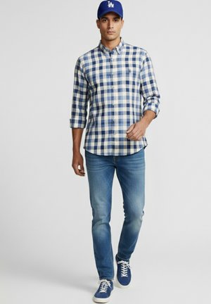 Hombre caminando con camisa de cuadros azul y blanca, vaqueros azules, zapatillas azules y una gorra de béisbol azul.