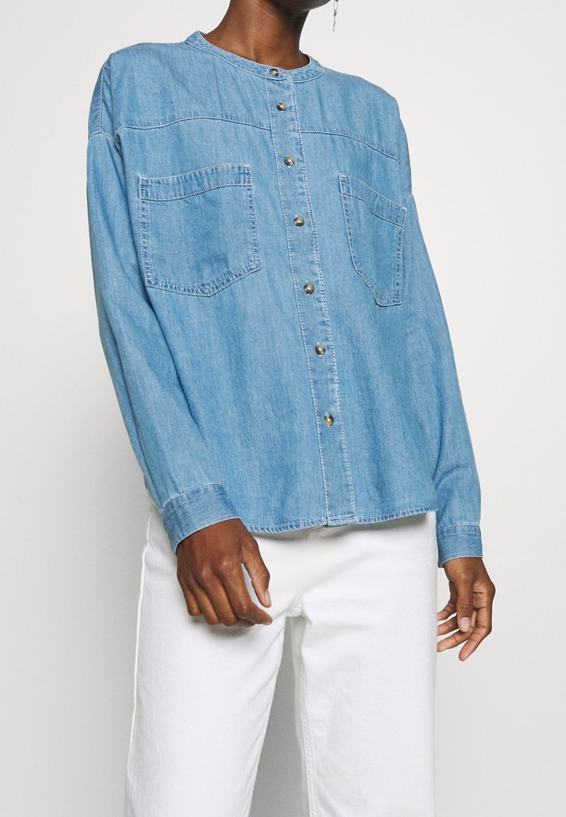Camicia in denim azzurro chiaro con bottoni, maniche lunghe, due tasche sul petto e scollatura rotonda, con bottoni argentati e una vestibilità morbida.