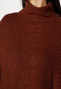 Brown, knitted turtleneck sweater with a ribbed texture. Features a high collar and vertical stitching detail across the fabric.