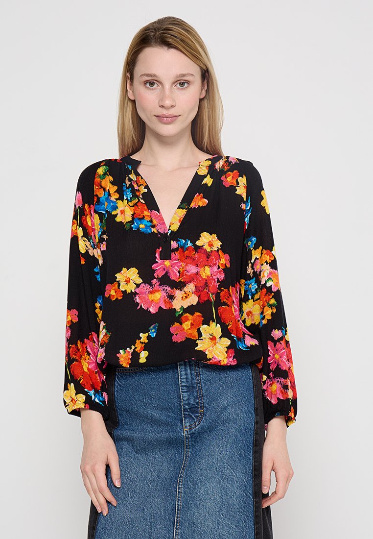 Kobieta z prostymi blond włosami nosi czarną bluzkę w kwiaty w czerwone, żółte i niebieskie kwiaty oraz dżinsową spódnicę, stoi przodem na tle białym.