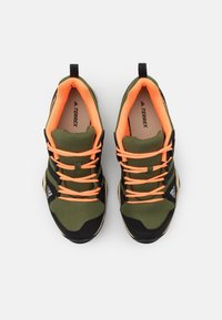 Scarpe sportive verdi e nere con lacci arancioni, parte superiore in rete e suola in gomma. Branding visibile sui lati e sulla linguetta. Design della punta arrotondata.