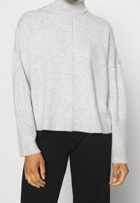 Maglione a collo alto lavorato a maglia di colore grigio chiaro, con bordi a costine. Vestibilità ampia e spalle cadenti, con una cucitura verticale lungo la parte frontale.