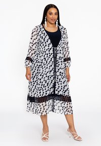 Leichtes, durchsichtiges weißes Kleid mit einem schwarz-braunen Punktmuster, elastischem Taillenbund, marineblauen Kontrast und 3/4 Ärmeln. Dazu kombiniert mit weißen Sandalen.