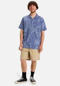 Kurzärmliges Hemd in Blau mit weißem Korallenmuster, kombiniert mit beigen Shorts. Dunkle Socken und schwarze Sneakers runden das Outfit ab.