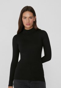 Schwarzes Langarmoberteil mit hohem Kragen, aus glattem Stoff. Figurbetontes Design mit Seitentaschen, kombiniert mit dunklen Jeans.