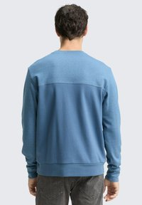 Niebieska bluza z kontrastowym, ciemniejszym niebieskim tyłem, długimi rękawami, dopasowanymi mankietami i okrągłym dekoltem. Miękki materiał o subtelnym wzorze.