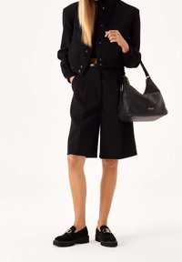 Giacca nera corta con chiusura a bottoni, abbinata a pantaloni shorts neri su misura. Gli accessori includono una borsa nera e mocassini neri con dettagli dorati.