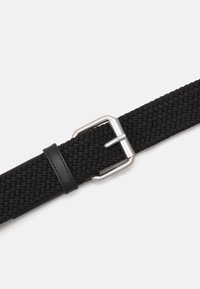 Cinturón negro tejido hecho de tela elástica con hebilla metálica plateada y acento de tira de cuero. Presenta un patrón texturizado en toda su superficie.