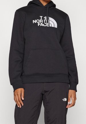 Persoon draagt een zwarte The North Face hoodie met voorzak en bijpassende zwarte broek met logo op het bovenbeen.