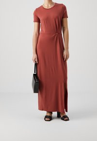 Rdzawoczerwona, krótko rękawna sukienka maxi z detalem w formie węzła w pasie, bocznym rozcięciem oraz gładkim materiałem. W połączeniu z czarną torebką i sandałami.