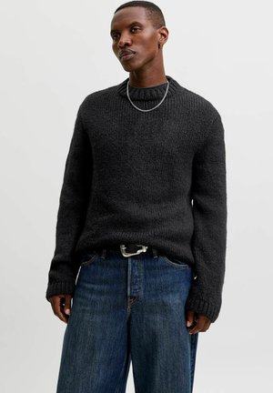 Homme portant un pull en maille gris foncé, un jean bleu à jambes larges avec une boucle de ceinture visible, et un collier chaîne argenté sur fond blanc.