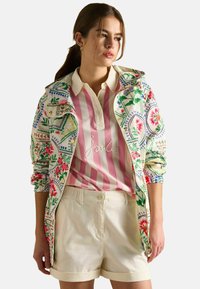 Jachetă ușoară cu imprimeu floral, albă, cu accente verzi și roz. Asortată cu un tricou polo cu dungi roz și pantaloni scurți bej.