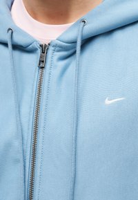 Κοντινό πλάνο ενός ατόμου που φοράει ανοιχτό μπλε φούτερ με φερμουάρ, με λευκό λογότυπο Nike και κορδόνια, πάνω από ένα απαλό ροζ πουκάμισο.