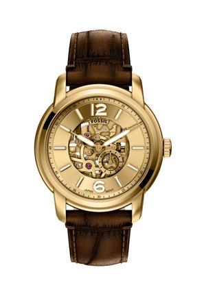 Montre de couleur or avec un cadran transparent, dévoilant le mouvement mécanique. Bracelet en cuir marron avec finition texturée. Présente de grands marqueurs d'heure.