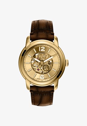 Montre de couleur or avec un cadran transparent, dévoilant le mouvement mécanique. Bracelet en cuir marron avec finition texturée. Présente de grands marqueurs d'heure.